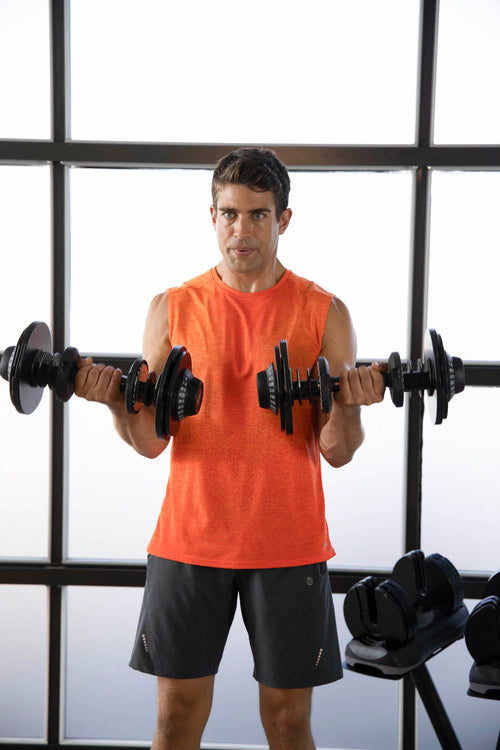 A man in a gym wearing an orange sleeveless shirt and dark gray shorts, lifting a barbell with dumbbells in front of a large window.