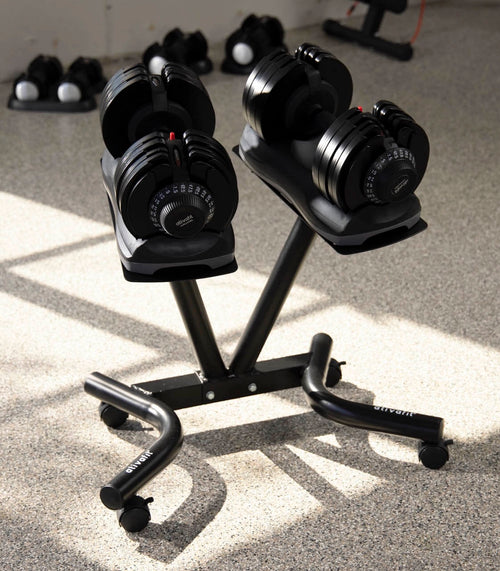 Black adjustable dumbbells on a matching black rolling storage stand, photographed in a real gym environment with floor shadows and other equipment blurred in the background.